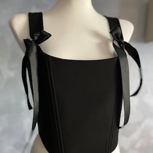 May include: A black corset top with adjustable straps that tie into bows. The corset has a square neckline and a fitted bodice.
