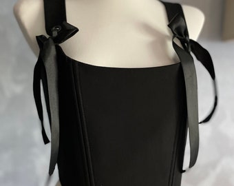 May include: A black corset top with adjustable straps that tie into bows. The corset has a square neckline and a fitted bodice.