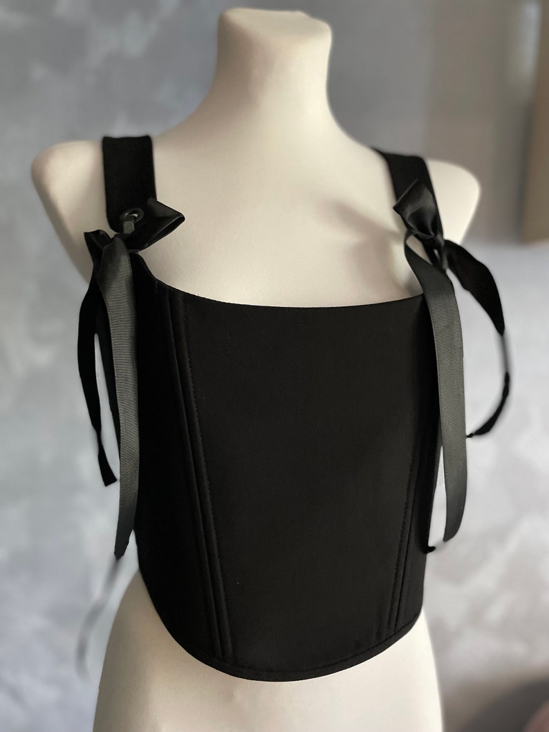 May include: A black corset top with adjustable straps that tie into bows. The corset has a square neckline and a fitted bodice.