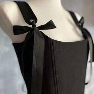 May include: A black corset top with adjustable straps and black ribbon ties. The straps have metal eyelets and the corset has a square neckline.