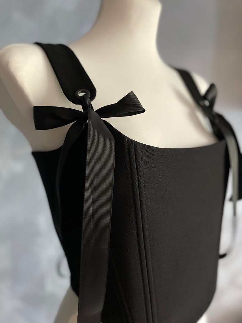May include: A black corset top with adjustable straps and black ribbon ties. The straps have metal eyelets and the corset has a square neckline.