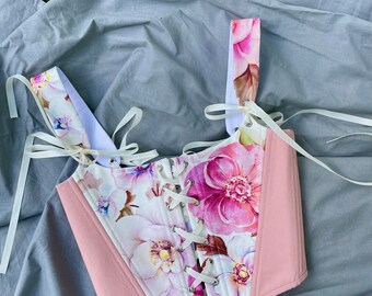 May include: A pink and white floral corset with white satin ribbon ties. The corset has a lace-up front and is made of a soft, comfortable fabric.