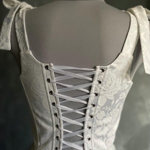 Op de afbeelding: Witte corset met een bloemenpatroon en kantversiering. Het corset heeft een gesnorte rug met wit lint.