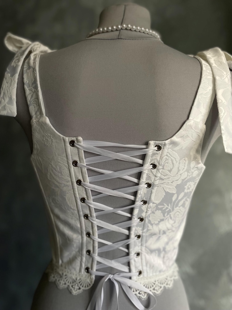 Op de afbeelding: Witte corset met een bloemenpatroon en kantversiering. Het corset heeft een gesnorte rug met wit lint.