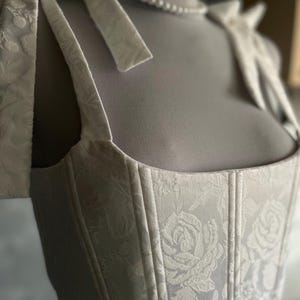 Op de afbeelding: Witte corset met een bloemenpatroon en baleinen. Het corset heeft een vierkante halslijn en verstelbare bandjes die aan de schouders worden vastgebonden.