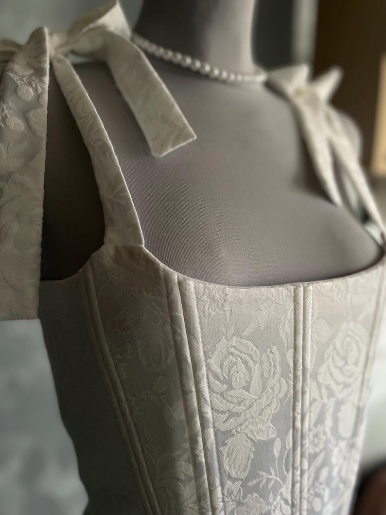 Op de afbeelding: Witte corset met een bloemenpatroon en baleinen. Het corset heeft een vierkante halslijn en verstelbare bandjes die aan de schouders worden vastgebonden.