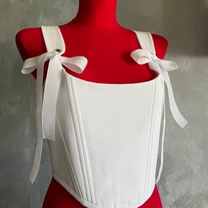 May include: White corset top with a square neckline and adjustable straps that tie into bows. The corset has a seam down the center front and two seams on each side.