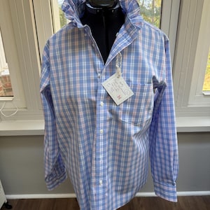 May include: A long-sleeved button-down shirt in a blue, white, and pink plaid pattern. The shirt features a prominent ruffled collar and a single chest pocket. A white rectangular tag is attached to the front, displaying handwritten text.