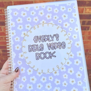 May include: A spiral-bound notebook with a light purple cover featuring white and gold floral patterns. The centre has a white circle with the text "Everly's Bible Verse Book" in black and gold lettering. The notebook is held against a brick wall.