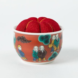 Pode incluir: Um alfineteiro vermelho numa tigela de cer&acirc;mica decorativa. A tigela laranja tem uma borda branca e apresenta uma cena pintada de figuras em trajes tradicionais. O alfineteiro &eacute; vermelho vibrante com costuras douradas.