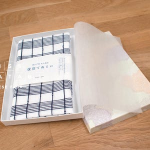 May include: An open white box displaying a folded Japanese fabric with a blue and white checked design. The box also contains a translucent paper cover. The words "MISS MATATABI" and "JAPANESE FABRIC" are visible on the box.