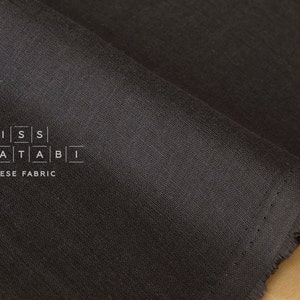 Puede incluir: Primer plano de una tela gris oscuro con una textura sutil. La tela est&aacute; doblada y tiene un borde sin rematar. El texto "MISS MATATABI JAPANESE FABRIC" est&aacute; impreso en la tela.
