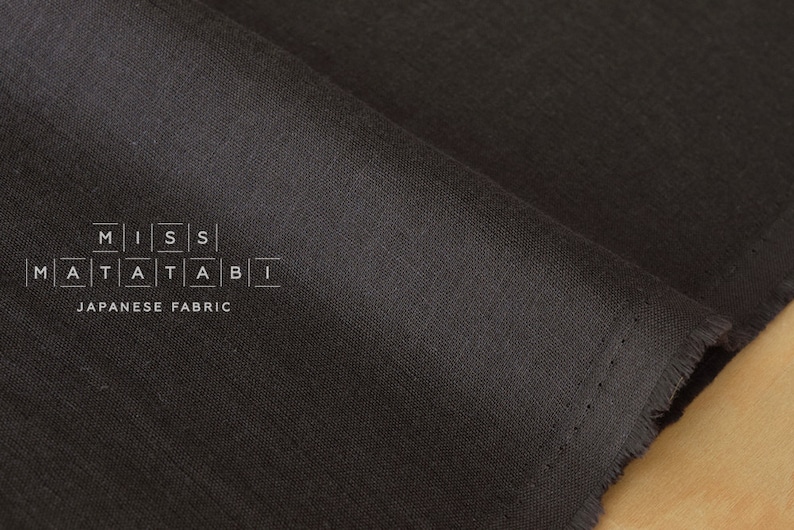 Puede incluir: Primer plano de una tela gris oscuro con una textura sutil. La tela est&aacute; doblada y tiene un borde sin rematar. El texto "MISS MATATABI JAPANESE FABRIC" est&aacute; impreso en la tela.