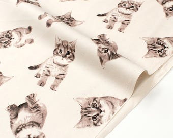 May include: Off-white fabric featuring a repeating pattern of adorable tabby kittens in various poses. The kittens are depicted in shades of brown and gray, with detailed fur and expressive eyes. The fabric is folded, showing its texture.