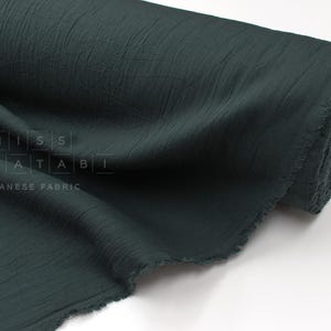 May include: A roll of dark forest green fabric with a crinkled, textured surface. The fabric is partially unrolled, showing soft folds and a raw, frayed edge. The text MISS MATATABI JAPANESE FABRIC is printed in white on the surface of the material.