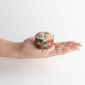 Pode incluir: Uma pequena tigela redonda de cer&acirc;mica vermelha e branca com um design decorativo de figuras, segurando um alfineteiro bege. A tigela &eacute; segurada na palma de uma m&atilde;o contra um fundo branco.