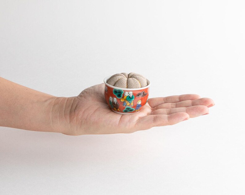 Pode incluir: Uma pequena tigela redonda de cer&acirc;mica vermelha e branca com um design decorativo de figuras, segurando um alfineteiro bege. A tigela &eacute; segurada na palma de uma m&atilde;o contra um fundo branco.