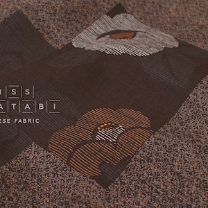 Puede incluir: Tela japonesa marrón con un estampado floral. La tela presenta un fondo texturizado con detalles en marrón oscuro y beige. El diseño incluye flores estilizadas en tonos marrón, beige y blanco. El texto "MISS MATATABI JAPANESE FABRIC" es visible.