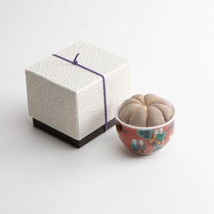 Pode incluir: Uma caixa de presente branca texturizada com um cord&atilde;o roxo, ao lado de uma pequena tigela decorada. A tigela &eacute; vermelha com um desenho pintado e cont&eacute;m uma almofada bege segmentada.