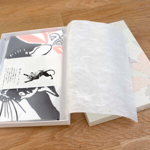 May include: A white gift box with a black and white cat design on the front. The box is open and the tissue paper inside is visible. The box is sitting on a wooden surface.
