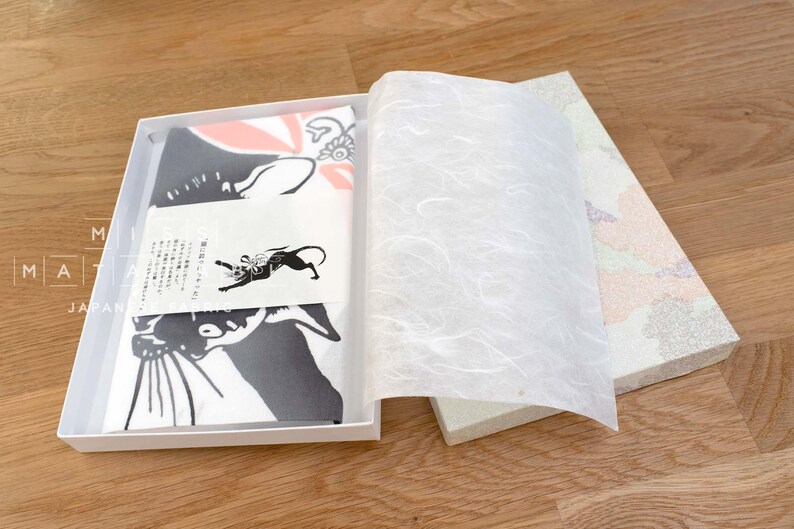 May include: A white gift box with a black and white cat design on the front. The box is open and the tissue paper inside is visible. The box is sitting on a wooden surface.
