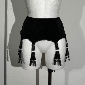 May include: Black vintage garter belt displayed on a white mannequin. The garter belt has a high waist and features six adjustable straps with metal clasps. The fabric appears to be a textured, matte material.