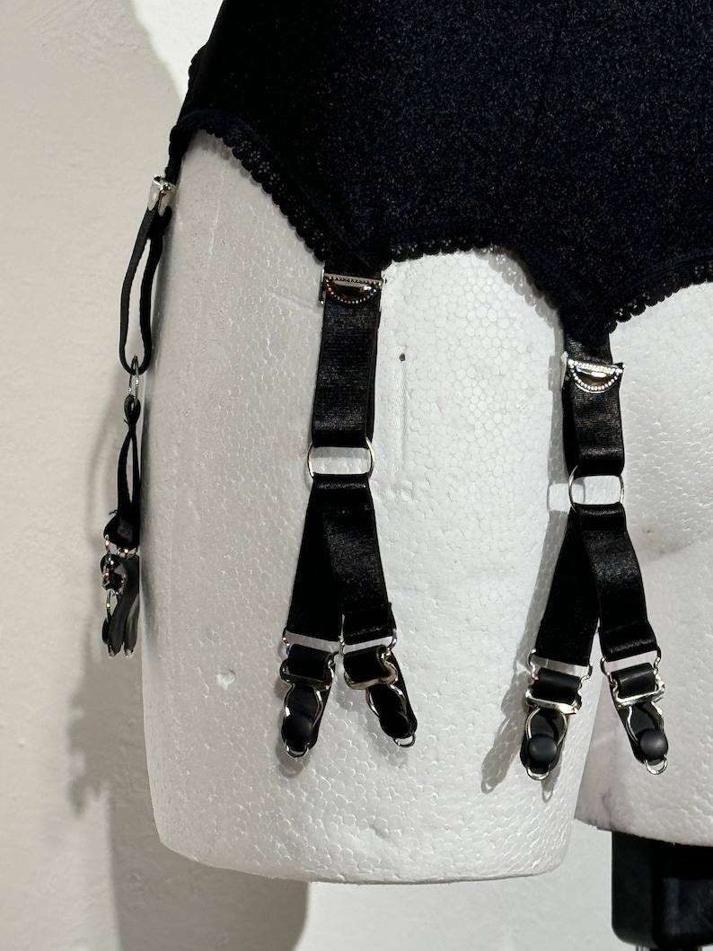 May include: Black garter belt with four adjustable straps and metal clasps. The belt is made of a textured fabric with a scalloped edge. The straps are made of a shiny black material.
