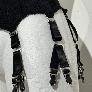 May include: Black garter belt with four suspender straps. The suspender straps have metal clips and adjusters. The garter belt is made of a black, textured fabric with a scalloped edge.