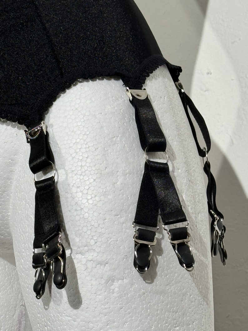 May include: Black garter belt with four suspender straps. The suspender straps have metal clips and adjusters. The garter belt is made of a black, textured fabric with a scalloped edge.