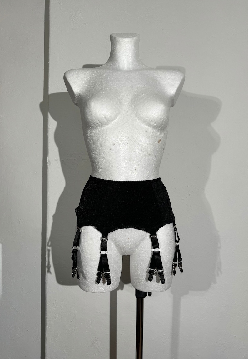 May include: Black vintage garter belt displayed on a white mannequin. The garter belt has a high waist and features six adjustable straps with metal clasps. The fabric appears to be a textured, matte material.