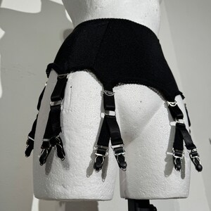 May include: Black garter belt with six straps and metal clasps. The belt is displayed on a white mannequin. The straps are made of black satin and have metal clasps to attach to stockings. The belt is made of a textured black fabric.