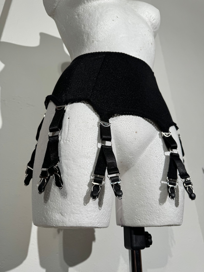 May include: Black garter belt with six straps and metal clasps. The belt is displayed on a white mannequin. The straps are made of black satin and have metal clasps to attach to stockings. The belt is made of a textured black fabric.