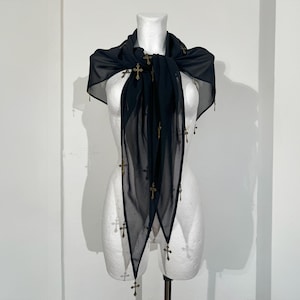 Könnte beinhalten: Ein schwarzer, durchsichtiger Schal mit goldfarbenen Kreuzanhängern. Der Schal ist über einer Schaufensterpuppe drapiert, was sein fließendes Design hervorhebt. Die Kreuze sind gleichmäßig verteilt und verleihen dem Schal ein dekoratives Element.