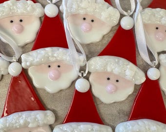 May include: A collection of Christmas ornaments shaped like Santa Claus faces. Each ornament features a red hat, white beard, and a pink nose. They are strung with white ribbons for hanging. The ornaments are made of glass.