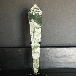 May include: A green and white moss agate crystal point on a black metal stand. The crystal is pointed at the top and has a smooth, polished surface.