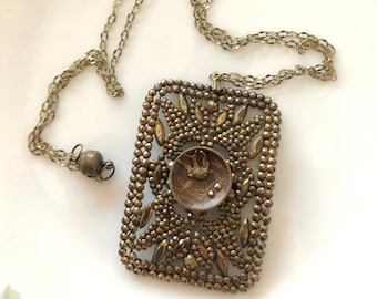 May include: A gold chain necklace with a rectangular pendant. The pendant is made of small metal beads and features a circular design with two birds in the center.