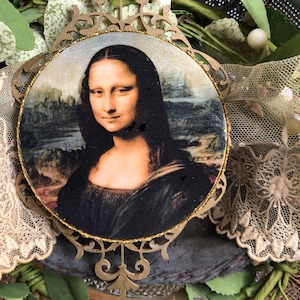 May include: A round, gold-framed ornament featuring a reproduction of the Mona Lisa painting. The ornament is decorated with a gold glitter border and has a detailed, ornate design.