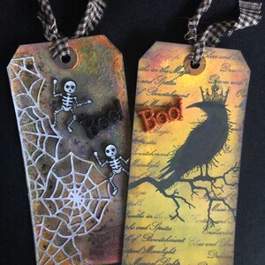 May include: Two Halloween-themed gift tags with black and white skeleton figures, a black crow, and the word "Boo" in orange glitter. The tags are decorated with a white spiderweb design and have a distressed, vintage look.