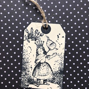 May include: A black and white illustration of a young girl wearing a crown and holding a stick with a ball on the end. The illustration is printed on a cream-colored tag with a black hole punched in the top.