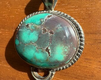May include: A silver pendant with a large oval turquoise stone. The stone has a green and brown marbled pattern. The pendant has a decorative bail with a rope-like design.