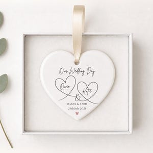 May include: A white heart-shaped ceramic ornament with a beige ribbon, featuring the black printed text Our Wedding Day, the names Owen and Katie inside heart outlines, the text BARNS & YARD, the date 25th July 2026, and a small pink heart.