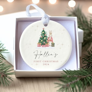 Puede incluir: Adorno navideño redondo y blanquecino con una ilustración en acuarela de un árbol de Navidad, un conejo y regalos. El texto "Hallie's First Christmas 2024" está escrito en una elegante escritura. El adorno está en una caja de regalo blanca.