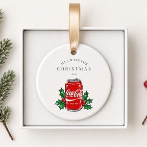 May include: A round white ceramic ornament featuring a red Coca-Cola soda can illustration surrounded by green holly leaves and red berries. The text above the can reads All I Want For Christmas is a. A gold ribbon is attached to the top.