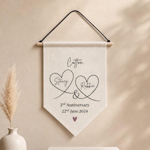 Könnte beinhalten: Ein beiges Baumwollbanner mit schwarzem Text und herzförmigen Designs. Das Banner lautet "Cotton Stacey & Robbie 2nd Anniversary 22nd June 2024" mit einem kleinen lila Herzen. Das Banner hängt an einem Holzstab.