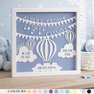 May include: A framed, layered paper art piece with a light blue background. White cut-out designs include hot air balloons, clouds, and the names "ALBERT GEORGE CASSIDY." Details include birth weight 8 lb 11 oz, time 08:38 PM, and date 08.09.2024.