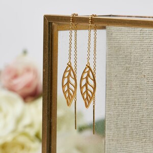 May include: Gold leaf earrings with delicate chain drop. The earrings are displayed on a gold stand against a white background.