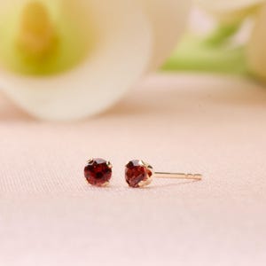 May include: A pair of stud earrings featuring round, deep red gemstones set in gold. The earrings are displayed on a soft pink surface, with a blurred white flower in the background. The jewelry is delicate and elegant.