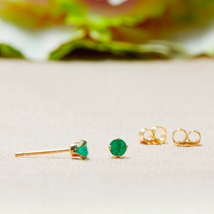May include: A pair of gold stud earrings with round, vibrant green gemstones. The earrings are displayed with two gold earring backs on a light pink surface. The earrings are a classic jewelry item.
