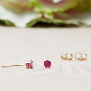 May include: A pair of gold stud earrings with round, bright pink gemstones. The earrings are displayed on a light pink surface, with two gold earring backs visible. The background features a blurred floral element.
