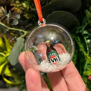 Puede incluir: Una bola de Navidad transparente con una botella de cerveza en miniatura en su interior. La botella es marrón con una etiqueta verde y blanca que dice "VICTORIA VB". La bola está llena de nieve artificial y tiene una cinta roja para colgar.